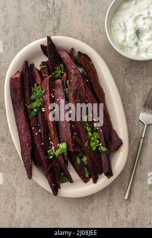 Fette di patate dolci viola al forno con spezie e prezzemolo in un piatto ovale di ceramica, salsa di avocado con yogurt e spezie Foto Stock