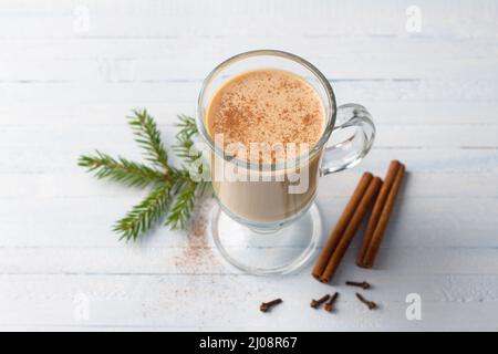 Tè piccante con latte in una tazza di vetro con cannella, biscotto di pan di zenzero legato con nastro e rami di albero di Natale su sfondo azzurro chiaro Foto Stock
