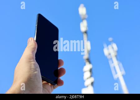 Smartphone in mano maschile sullo sfondo delle torri cellulari. Apparecchiature per telecomunicazioni mobili, potenza del segnale telefonico Foto Stock