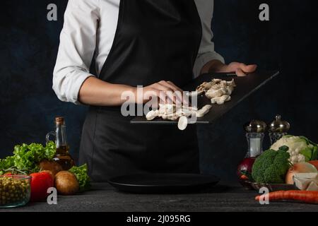 Lo chef professionista in grembiule nero versa i funghi tritati sul piatto per cucinare la zuppa su sfondo blu scuro. Backstage di preparazione del pasto. Healt Foto Stock