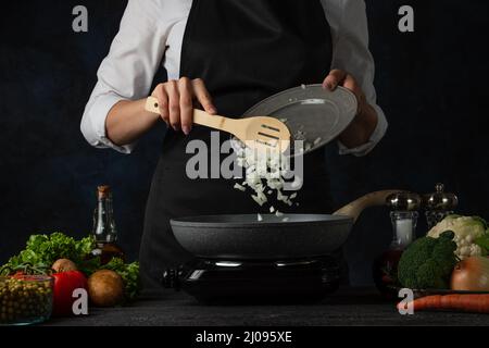 Lo chef professionista in grembiule nero versa la cipolla tritata in una padella per cucinare la zuppa su sfondo blu scuro. Backstage di preparazione del pasto. Di. Sano Foto Stock