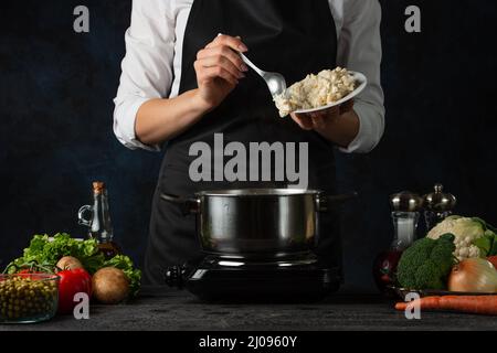 Lo chef professionista in grembiule nero versa il formaggio tritato in una padella per cucinare la zuppa su sfondo blu scuro. Backstage di preparazione del pasto. Piatto sano. FO Foto Stock