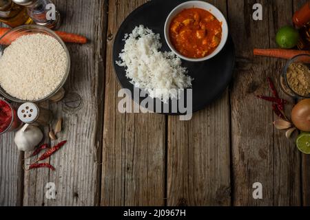 Vista dall'alto di un piatto rotondo nero con riso bollito e gustoso curry di pollo caldo su un rustico tavolo di legno con fondo di lime e spezie. Indi. Classico Foto Stock