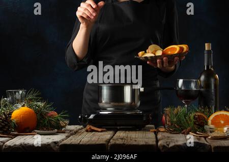 Lo chef professionista versa la frutta in una padella per preparare il VIN brulé su un rustico tavolo in legno con fondo di composizione festiva. Backstage di cottura caldo Foto Stock