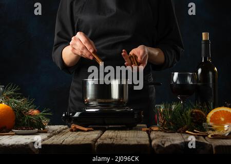 Lo chef professionista versa il bastone di cannella in una padella per preparare il VIN brulé su un rustico tavolo di legno con fondo di composizione festiva. Backstage di coo Foto Stock