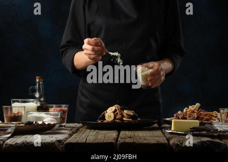 Lo chef professionista prepara fiocchi di cocco per servire le cialde su un rustico tavolo di legno con ingredienti su sfondo blu scuro. Backstage di cottura s Foto Stock