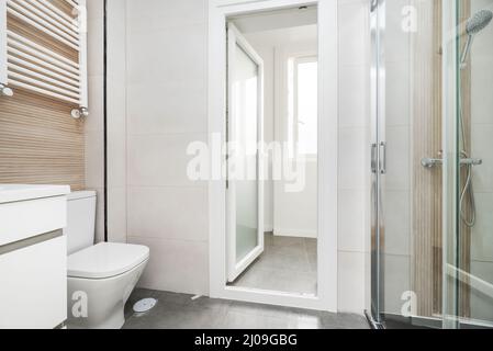 wc piastrellato con grandi piastrelle bianche, lavandino in porcellana bianca, radiatore in alluminio bianco e box doccia con divisorio in vetro Foto Stock