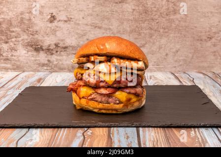Hamburger di bestia con manzo doppio, formaggio cheddar doppio, funghi stufati, pancetta fritta e salsa barbecue su piatto di ardesia nera su tavola di legno Foto Stock