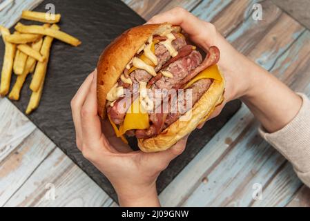 Le mani della persona tengono un hamburger di carne di tre bestie con un sacco di pancetta fritta, formaggio cheddar e salsa per darvi un attacco di cuore per dargli una buona punta Foto Stock