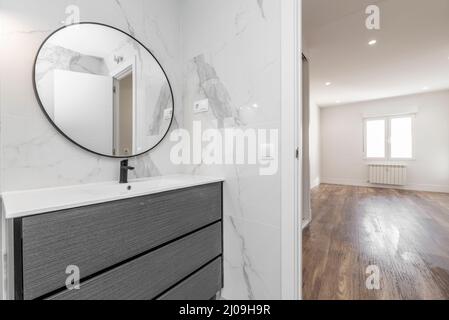 Bagno con lavandino in porcellana su comò grigio, specchio circolare con cornice nera e accesso alla camera da letto vuota con pavimenti in legno Foto Stock