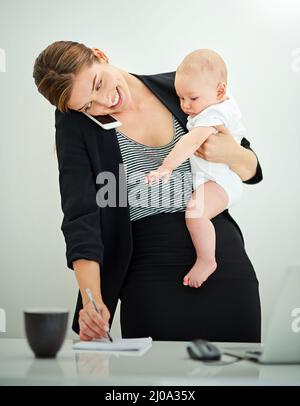 Sollevare un business e un bambino. Colpo di una giovane donna d'affari riuscita che trasporta il suo bambino adorabile mentre parla al telefono. Foto Stock