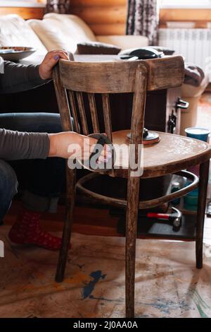 Lady lentamente rimuovendo il vecchio strato di vernice levigando parte posteriore di sedia antica con carta vetrata in officina per la ristrutturazione dei mobili. Pratiche sostenibili Foto Stock