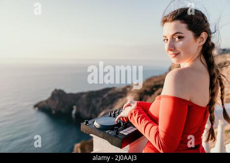 Le mani femminili del DJ suonano la musica mescolando e graffiando durante la festa serale con sfondo marino e caldo tramonto morbido. Primo piano di una console DJ controllata da Foto Stock