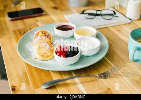 Sana colazione estiva. Pancake al formaggio casolare con frutti di bosco freschi e cappuccino su tavola di legno. Primo piano Foto Stock