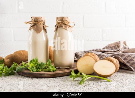 Il latte vegetale vegano di patate è una nuova tendenza nel consumo di cibo: Due bottiglie di latte di fronte ad un muro di mattoni bianchi su un vassoio di legno Foto Stock