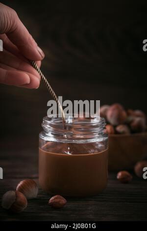 Latte di nocciole di cioccolato fatto in casa spalmato su vaso di vetro su sfondo di legno scuro. La mano della donna tiene un cucchiaio Foto Stock