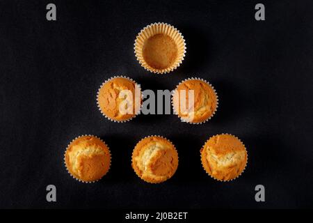 Still LIFE con vista dall'alto di un gruppo di cupcake con uno dei contenitori vuoto, posto in forma triangolare, su sfondo scuro. Foto Stock