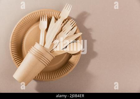 Vasellame di carta monouso. Tazze, piatti, forcelle in legno, coltelli e cucchiai su sfondo beige. Senza plastica e senza rifiuti. Foto Stock