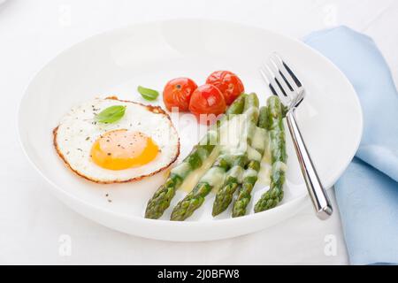 Uovo fritto con asparagi e pomodori su un piatto bianco con tovagliolo blu. Foto Stock