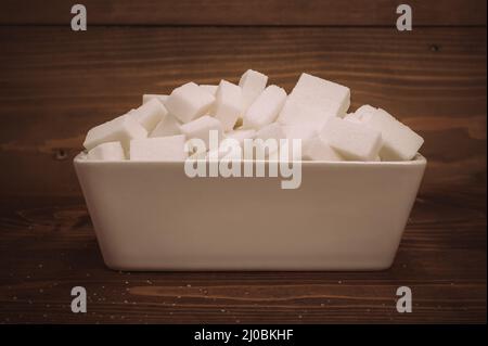 Cubetti di zucchero bianco in ciotola di ceramica su sfondo di legno. Primo piano Foto Stock