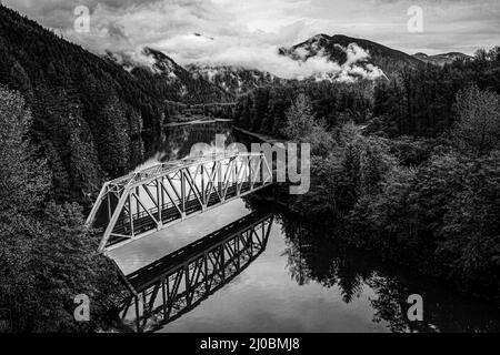 Scala di grigi aerea di un ponte di metallo sul fiume circondato da foresta sotto le nuvole Foto Stock