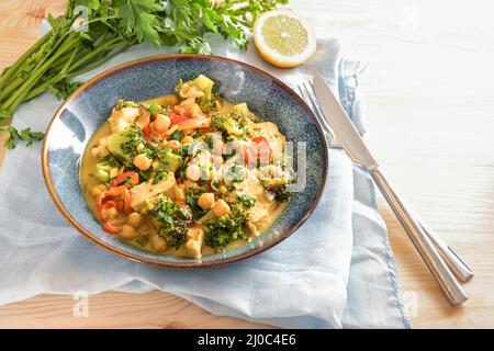 Piatto di curry da ceci, broccoli, peperone e carne di pollo su un piatto blu, servito con posate, erbe e tovagliolo su un tavolo di legno chiaro, copia Foto Stock