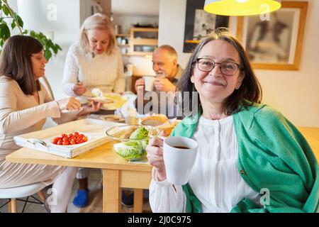 Donna anziana che ha una tazza di caffè in cucina con gli amici che hanno la colazione in background Foto Stock