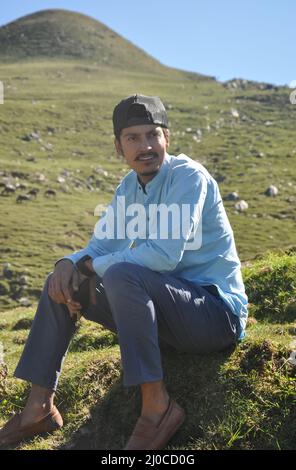 Un giovane indiano bello osservare seduto sull'erba in montagna con guardando lateralmente Foto Stock