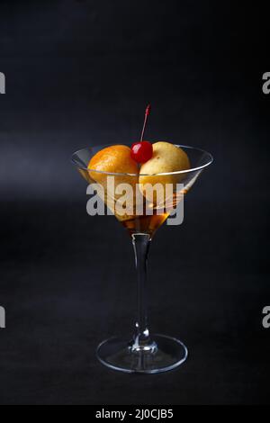 Rum baba napoletano (o baba au rum) in un bicchiere di martini con una ciliegina da cocktail su sfondo nero. Piccole torte di lievito imbevute di sciroppo di rum. Tradizione Foto Stock