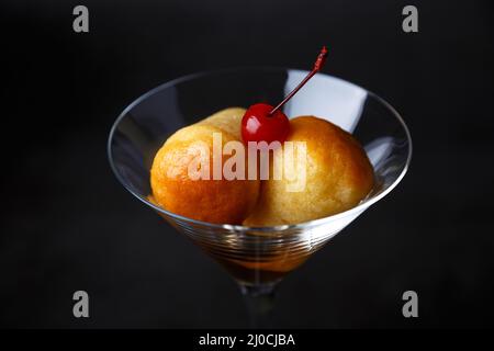 Rum baba napoletano (o baba au rum) in un bicchiere di martini con una ciliegina da cocktail su sfondo nero. Piccole torte di lievito imbevute di sciroppo di rum. Tradizione Foto Stock