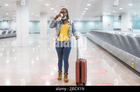 Donna africana che cammina nel terminal dell'aeroporto con i bagagli mentre beve una tazza di caffè Foto Stock