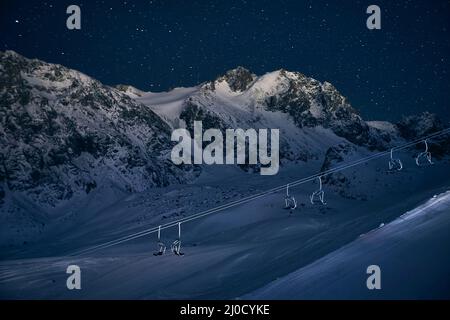 Funivia vuota e pista da sci al parco notturno con alta montagna contro il cielo stellato presso la stazione sciistica di Shymbulak Almaty, Kazakhstan Foto Stock