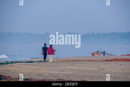 Sindhudurg, India - 20 dicembre 2021 : Coppia non identificata che prende una passeggiata mattutina sul lato della spiaggia con uccelli gabbiano che volano e barca da pesca nel bac Foto Stock