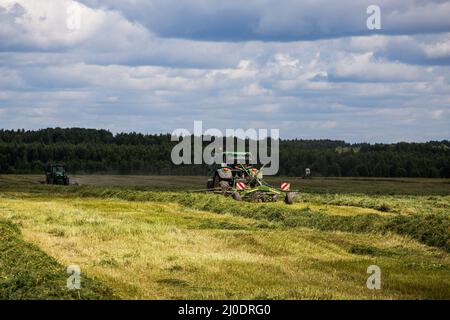 Due trattori John Deere verdi haymaking con aratro Krone sul campo estivo prima della tempesta - teleobiettivo con fuoco selettivo Foto Stock