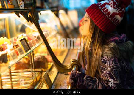 Un bambino nei vestiti di inverno e un cappello rosso di Natale sceglie i dolci in una finestra del negozio per comprare. Il concetto di Capodanno vacanze, sconti, shopping. Foto Stock