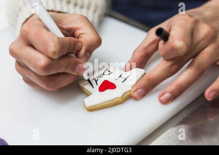Vista ravvicinata di una donna pasticcera professionista che scrive e disegna a mano un biscotto. Foto Stock