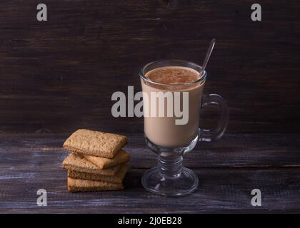 Tè piccante con latte in una tazza di vetro con cannella, biscotti di pan di zenzero su un tavolo di legno, stile rustico Foto Stock