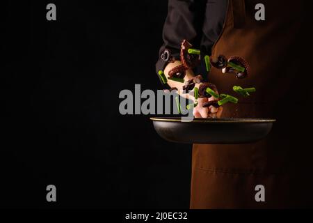 Uno chef professionista prepara polpi con pezzi di verdure in una padella su sfondo nero. Pesce in volo congelato. Cibo sano, sano Foto Stock