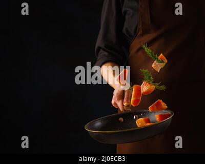 Uno chef professionista prepara pezzi di trota rossa, salmone con verdure in una padella. Frutti di mare in volo congelato su sfondo nero. Ristorante, hotel, Foto Stock