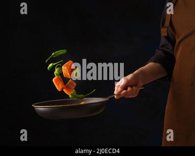 Uno chef professionista su sfondo nero prepara pezzi di pesce rosso con verdure in una padella. Levitazione. Cibo vegetariano sano, ristorante Foto Stock