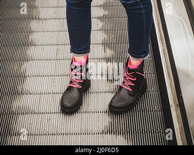 Le gambe femminili in jeans blu e gli stivali in gomma nera bagnata con lacci rosa si trovano sulla scala mobile Foto Stock