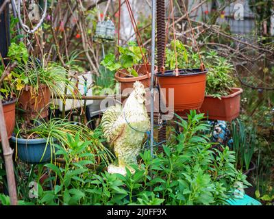 Una statuina ceramica da giardino di un gallo si trova nel giardino tra vasi di fiori appesi e in piedi con piante Foto Stock