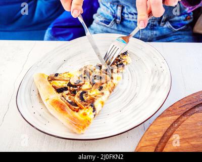 Le dita stanno tenendo una forcella e un coltello mentre affettano a. pezzo di pizza di funghi che giace su un piatto Foto Stock
