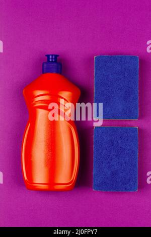 Bottiglia di plastica arancione di liquido per lavastoviglie e spugna blu su sfondo viola, vista dall'alto ravvicinata Foto Stock