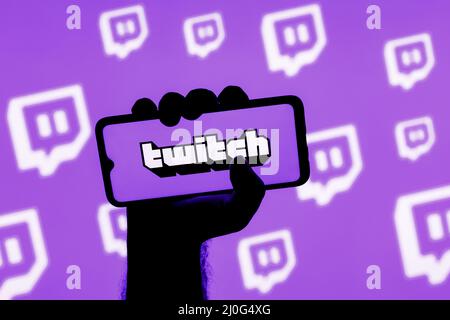 Smartphone con il logo del servizio di streaming live Twitch sullo schermo in una mano appesa sullo sfondo dei logo Twitch Foto Stock