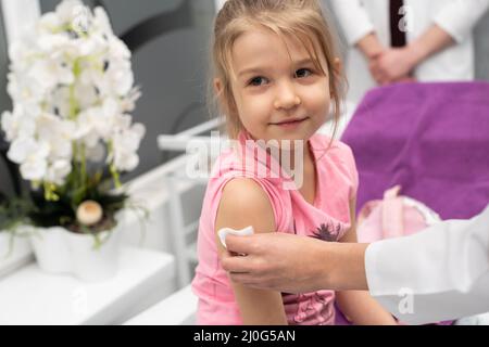 La ragazza sta guardando fuori dalla cornice. Una giovane donna medico sta preparando un bambino per un'iniezione disinfettando un pezzo di pelle Foto Stock