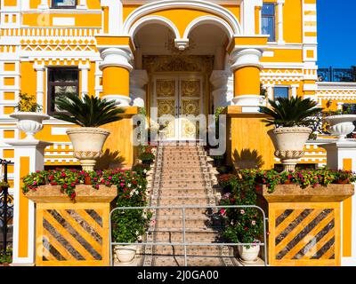 Frammento di un edificio giallo con ornamento bianco, colonne, porte con motivo dorato e gradini ricoperti di moquette Foto Stock