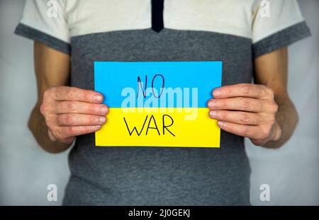 Un uomo in piedi su un grigio, tiene in mano lo sfondo di un cartoncino giallo-blu bandiera Ucraina con l'iscrizione 'No war' Foto Stock