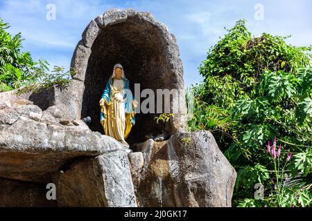 Statua della Vergine Maria in una cappella rupestre fuori dalla chiesa cattolica di Sant’Anna a Baie Ste Anne, Praslin, Seychelles, con vegetazione tropicale Foto Stock