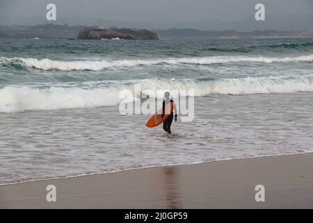 Un surfista che cammina in mare portando la sua tavola da surf con il faro sull'isola di Moors in lontananza Sardinero spiaggia Santander Cantabria Spagna Foto Stock